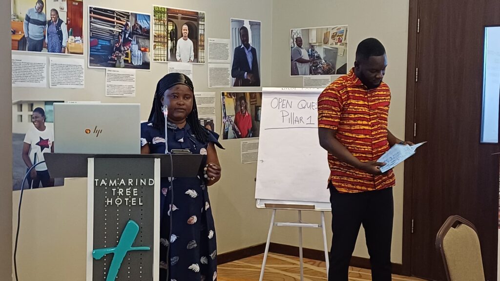 Two people are giving a presentation in a conference room. One is speaking at the podium with a laptop, and the other is holding a paper. A flip chart reads "Open Questions Pillar 2.