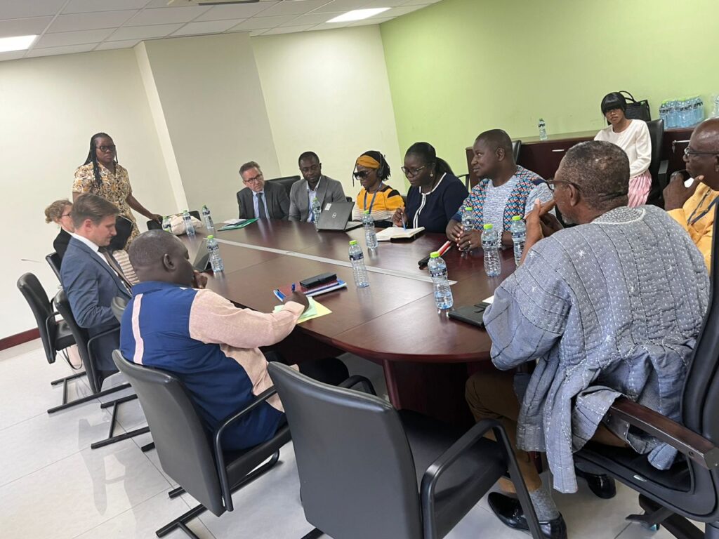 Photo description: The photo depicts a meeting with a group of twelve people, including Members of Parliament, GFD Management, and UNCRPD Officials, gathered around a large, oval conference table in a meeting room with pale green walls. The participants, a mix of men and women of various ages and ethnic backgrounds, appear engaged in discussion, with some taking notes. Water bottles, notebooks, laptops, and pens are placed in front of most attendees. One woman stands, addressing the group, while others are seated and attentive.